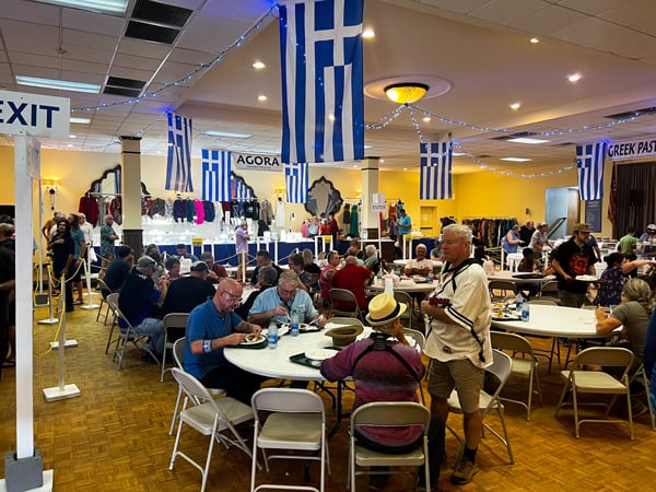 Asheville Greek Festival Agora Area with tables and chairs filled with people eating and talking surrounded by food and vendors with Greek flags hanging as decor