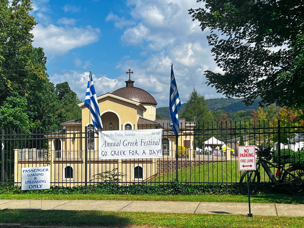Asheville Greek Festival entrance with fence with signs, Greek flags, and Holy Trinity Greek Orthodox Church of Asheville in the background