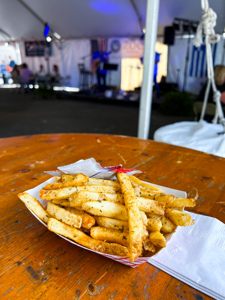 Asheville Greek Festival Greek Fries with seasoning in plastic holder on brown table with Greek band playing music in the background