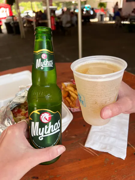 Asheville Greek Festival Mythos Beer and local craft cider with two white hands holding each and doing a cheers over a table with a falafel in the background