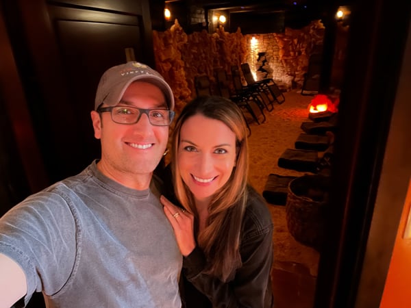Tom and Christine, a white brunette male and female, taking a selfie in front of community salt cave with orange lighting at the Asheville Salt Cave (North Carolina)