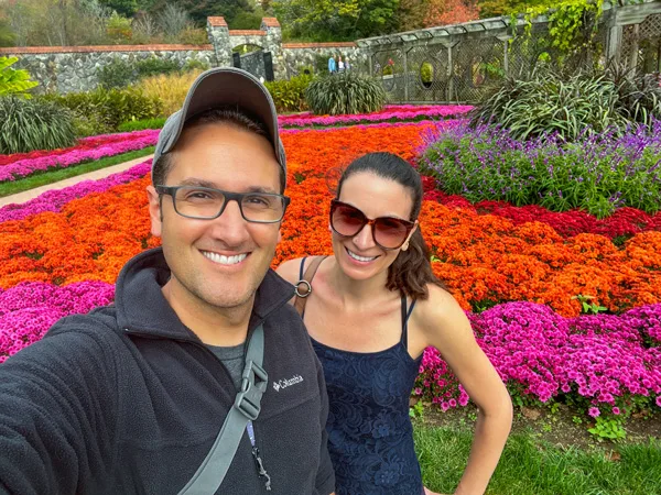 Biltmore Walled Gardens during the Fall in Asheville NC with white brunette male wearing hat, glasses, and fleece and white brunette female with sunglasses and tank taking a selfie in front of orange and pink flowers