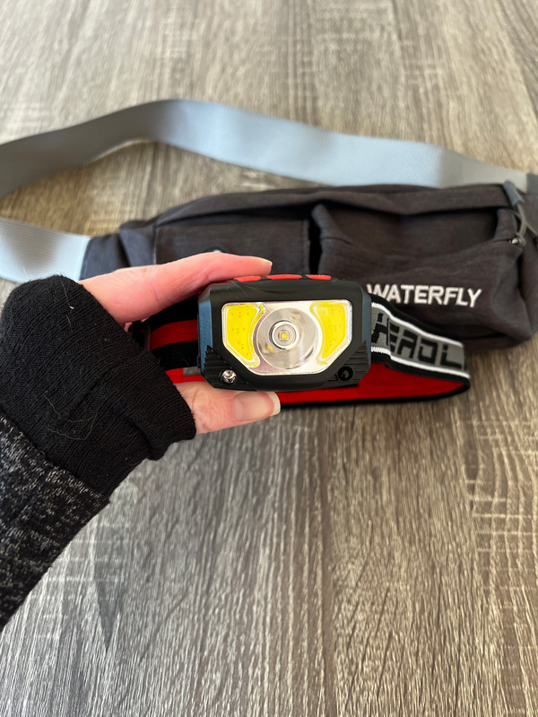 white hand in gray top holding up a headlamp over table with fanny pack on it