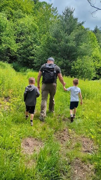 White brunette male holding two children's hands as they walk along a grassy dirt trail in hiking shorts and shirts