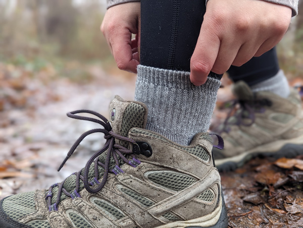 gray Merino Wool Socks with white hands pulling them up and tan hiking boot with purple laces