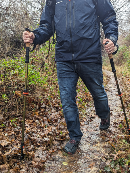 white person in blue rain jacket and jeans with Trekking Poles on a muddy dirt trail outside