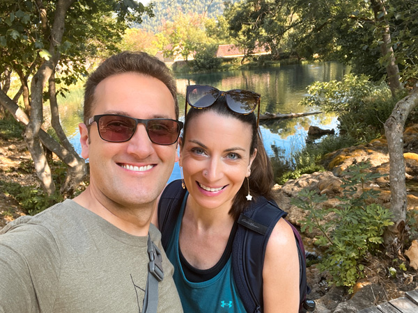 White brunette and female with sunglasses, taking a selfie outside in front of small body of water