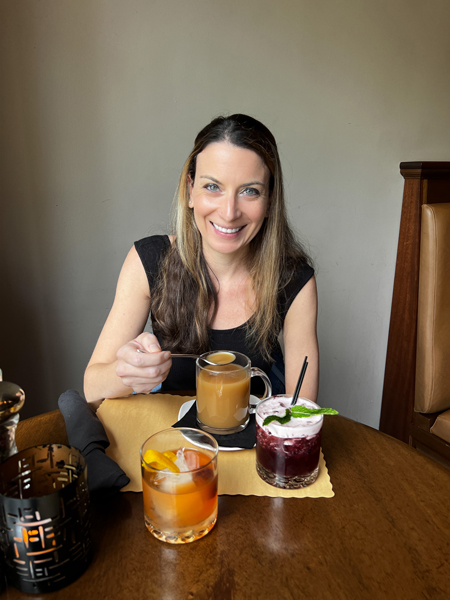 Christine, a white brunette female drinking Bone Broth from a clear glass with two cocktails on the table at Asheville restaurant, Bone and Broth