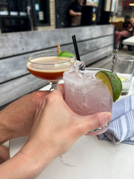 Two white hands doing a cheers with two cocktails, one pink and one brown. The pink drink is a Zero Proof Cocktail. The restaurant is Crave Dessert Bar in Asheville 