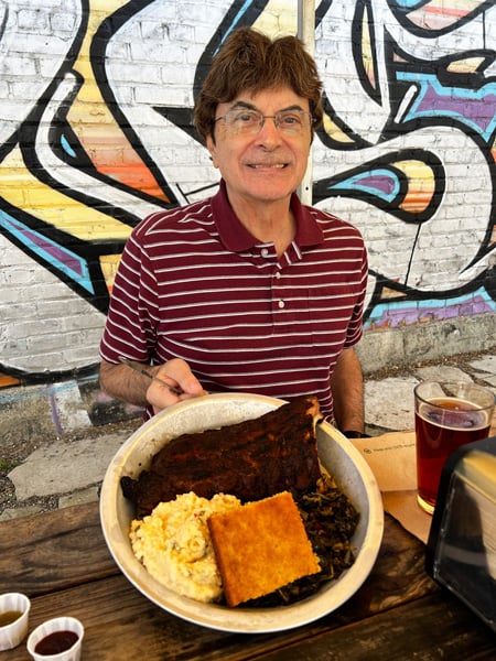 Christine's dad, a white brunette male in red striped shirt eating a BBQ rib platter with corn bread and potato salad at 12 Bones Smokehouse River location in Asheville, NC