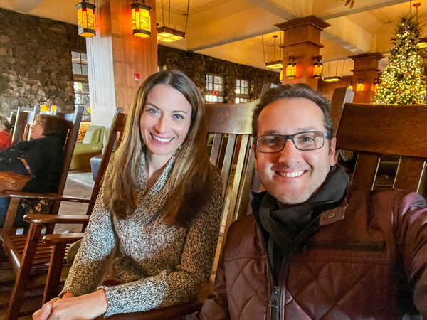 Christine and Tom, white brunette male and female in sweaters sitting in rocking chairs at The Omni Grove Park Inn in Asheville at Christmas with lighted tree in the background