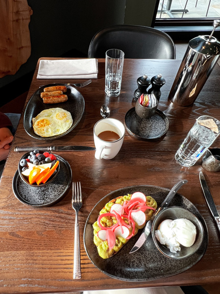 Breakfast at the Grand Bohemian Lodge Greenville at their on-site restaurant, Between the Trees with a plate of avocado toast and poached eggs, another plate of over easy eggs with sausage and fruit, and French Press with coffee mugs