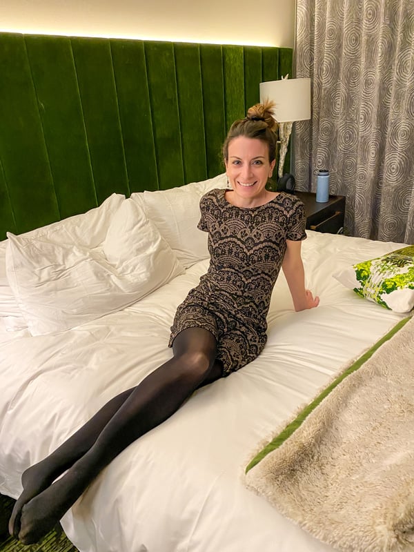 Christine, a white brunette female in a black and gold dress sitting on a white sheet bed at the Grand Bohemian Lodge Greenville