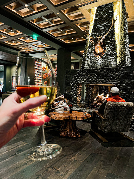 The lobby of the Grand Bohemian Lodge Greenville with a large stone fireplace with deer head taxidermy decoration and Christine's white hand holding up a glass of white wine with bookshelves in the background