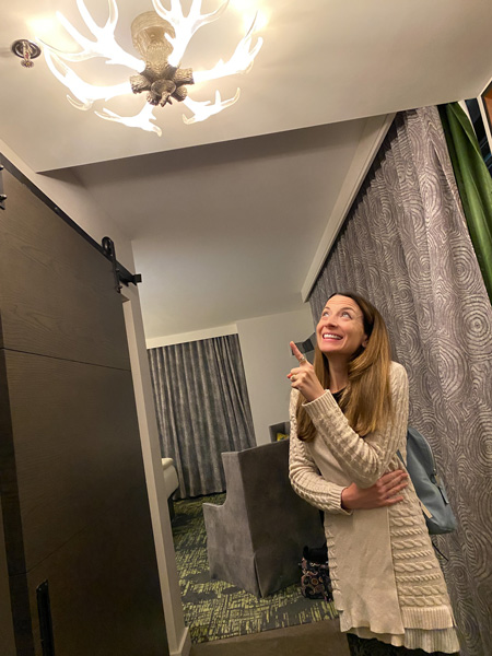 Christine, a white brunette female in a beige sweater, pointing at antler themed light in her hotel room at the Grand Bohemian Lodge in Greenville, SC