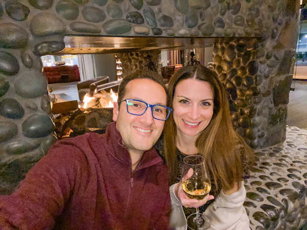 Christine and Tom, a white brunette male and female in winter sweaters, taking a selfie in front of the blazing stone fireplace at the Grand Bohemian Lodge Greenville (South Carolina)