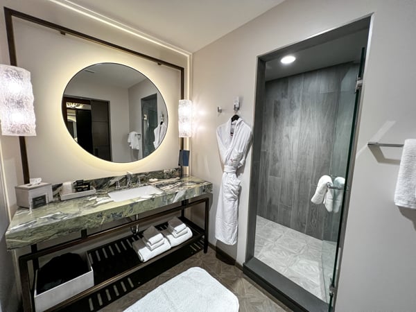 Our hotel guest room bathroom at the Grand Bohemian Lodge Greenville, which has a large round, lighted mirror over a green marble sink with a walk-in shower to the right and white bathroom hanging on the wall