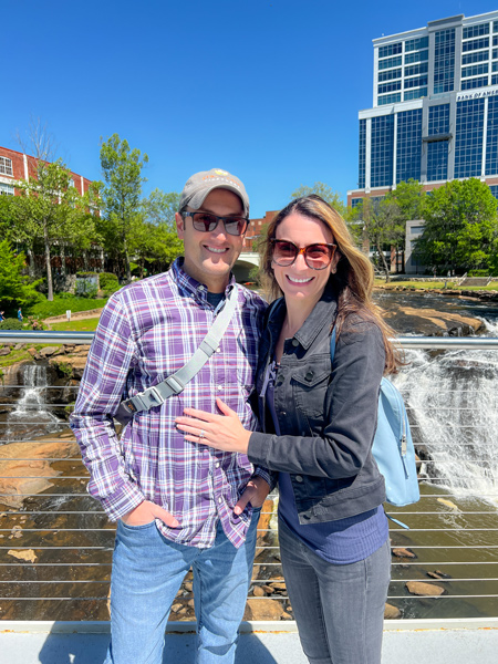 Tom and Christine, a white brunette male and female in fall clothes with sunglasses, getting their photo taken on Liberty Bridge (a suspension bridge over the water) at Falls Parks on the Reedy in Greenville, SC