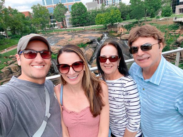 Selfie with Christine, Tom, and Christine's parents (four white, brunette people) on Liberty Bridge with falls and business buildings in the background in Greenville, SC