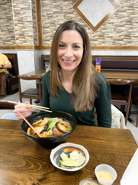 Christine, a white brunette female in a green sweater, eating a bowl of ramen at a brown table at Mizu Noodle and Asian Cuisine in Asheville, NC