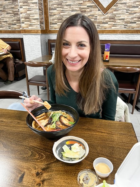 Christine, a white brunette female in a green sweater, eating a bowl of ramen at a brown table at Mizu Noodle and Asian Cuisine in Asheville, NC