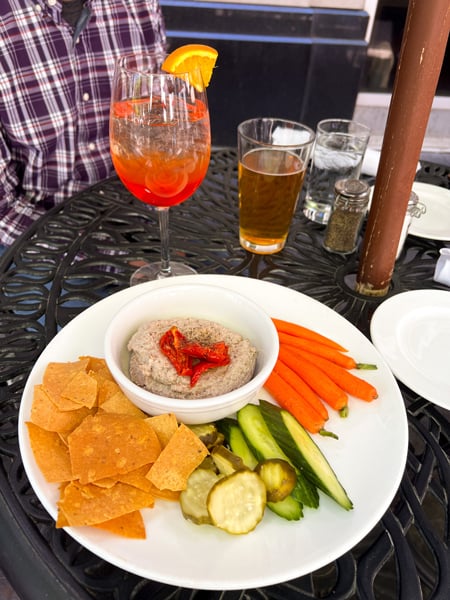 Hummus plate with carrots, cucumbers, and pickles at the Nose Dive Restaurant in Greenville, SC; Tom is in the background in a purple plaid shirt with an orange cocktail