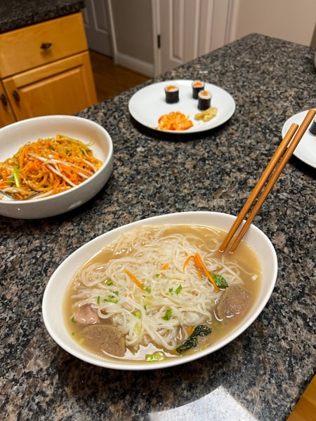Takeout pho soup (noodles, brown broth, meats, and carrots) in a white bowl on our kitchen island with chopsticks from Suwana's Asian Cuisine in Asheville, NC